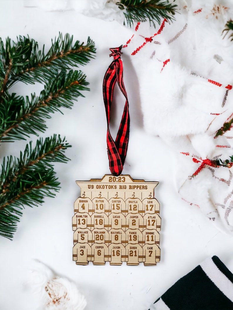 May include: A wooden hockey ornament with the text "20:23 U9 Okotoks Big Rippers" and the names and numbers of a hockey team. The ornament is brown and has a red and black checkered ribbon attached.