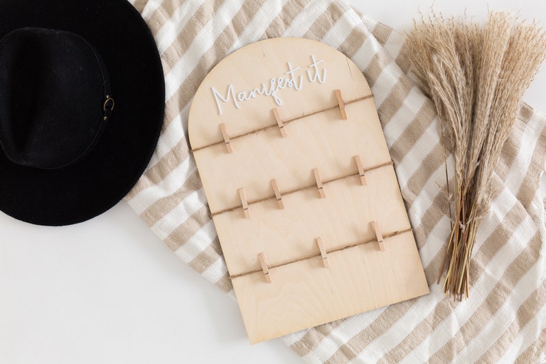 May include: A wooden board with the text "manifest it" written in white lettering. The board has 10 clothespins attached to it with twine.