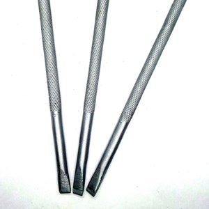 Puede incluir: Tres herramientas de metal plateado con mango texturizado y punta plana y puntiaguda. Las herramientas están dispuestas en forma de V.