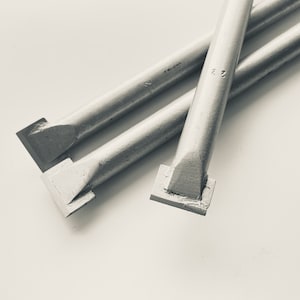 Puede incluir: Tres cinceles de metal plateado con cabezales cuadrados, sobre una superficie blanca.