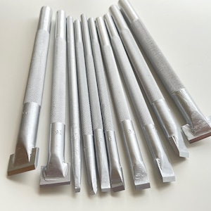 Puede incluir: Un conjunto de ocho herramientas de cuero de metal plateado con puntas de diferentes formas. Las herramientas tienen una superficie texturizada y están dispuestas en forma de abanico.