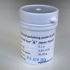 May include: A white plastic jar with a black label that reads "Diamond polishing paste 0,1/0 Diamond Tool" with a water drop icon. The label also includes the text "(Water Based)" and lists the size of the diamond particles as "0,1 μ", the carat weight as "4,00 carat", and the weight of the paste as "40,00 g". The label also includes the date "06 APR 2020" and the website "mpx90pin.com".