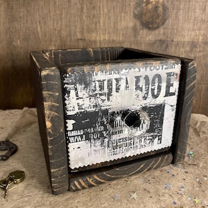 May include: A small wooden box with a black stain finish. The front of the box has a vintage-style paper label with the text "CUT A HOLE" printed on it.