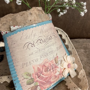 May include: A blue metal tin with a white label that reads "Early Morning Ballad with an Accompaniment for the Piano Forte Harp Composed by D. Kemp". The tin is decorated with a pink rose and a white lace ribbon.