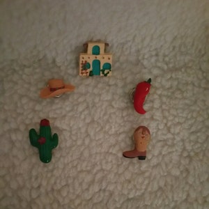 May include: Five small, colorful, ceramic figurines with a western theme. The figurines include a cactus, a cowboy boot, a red chili pepper, a cowboy hat, and a small adobe building.