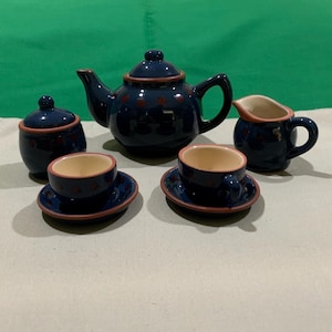May include: A miniature blue ceramic tea set with red heart accents. The set includes a teapot, sugar bowl, creamer, and two teacups with saucers. The rims are trimmed in red. The set is designed for children's play or as a decorative item.