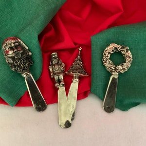May include: Set of four silver Christmas themed butter knives. The knives feature a Santa Claus head, a Christmas tree, a nutcracker, and a wreath.