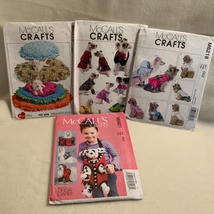 May include: Four McCall's Crafts sewing pattern packets. The packets feature images of dog clothing and accessories. One packet shows a child wearing a dog-themed apron. The packets are white with the McCall's Crafts logo.