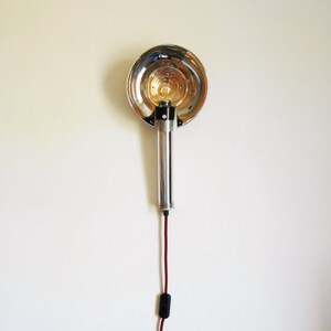 Peut inclure: Une lampe murale chromée avec un réflecteur rond et un cordon rouge. La lampe a un style vintage.