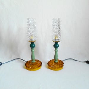 Może przedstawiać: Para lamp stołowych w stylu vintage. Każda lampa ma przezroczysty, teksturowany szklany klosz na zielonej i złotej podstawie. Podstawa ma okrągły, bursztynowy, teksturowany wzór. Lampy są podłączone do szarego przewodu zasilającego.