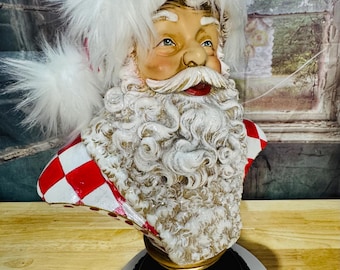 May include: A Santa Claus bust with a white fluffy hat and beard. The bust has a red and white checkered pattern on the shoulders. The base is dark with a gold accent and holly berry design. A festive holiday decoration.