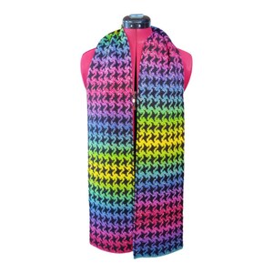 May include: A colorful scarf with a houndstooth pattern in a rainbow gradient. The scarf is draped over a pink mannequin, showcasing the vibrant hues of pink, purple, blue, green, and yellow. The scarf's design is a repeating pattern.