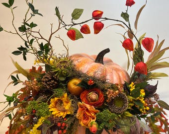 May include: A large orange pumpkin decorated with fall foliage, flowers, and berries. The pumpkin is surrounded by sunflowers, roses, and other autumnal blooms.
