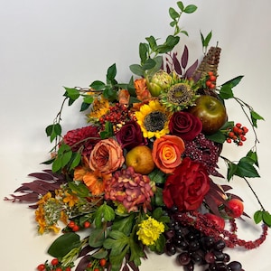 May include: A vibrant floral arrangement featuring sunflowers, roses, and other colorful blooms. The bouquet includes grapes, apples, and berries, creating a harvest-themed display. The arrangement is set against a white background.