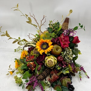 May include: A fall-themed cornucopia arrangement with a variety of artificial fruits, vegetables, and flowers. The arrangement features a sunflower, artichoke, apples, grapes, pine cones, and burgundy roses.