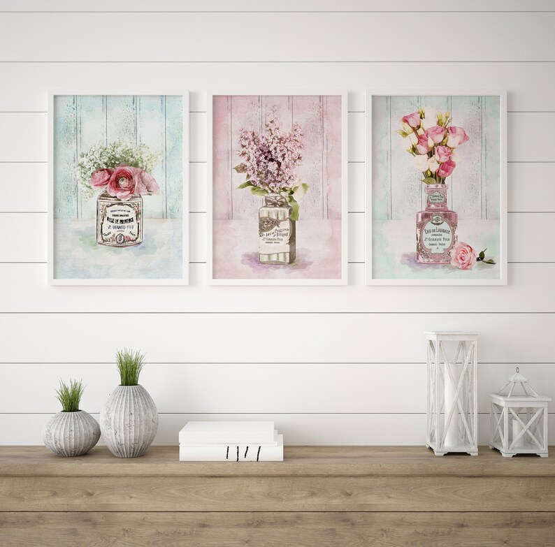 May include: Three framed watercolour prints of floral arrangements in vintage bottles. The prints feature pink and white flowers, and the bottles have labels with French text. The prints are displayed on a white shiplap wall with a wooden shelf below.