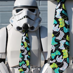 May include: A black tie featuring a pattern of white, green, and teal video game controllers. The tie is worn by a white Stormtrooper helmet and armor. The controllers are connected by dashed lines.