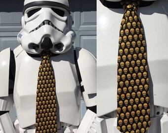 May include: A black necktie with a repeating pattern of yellow skulls. The tie is worn by a person in a white Stormtrooper costume. The tie is a unique accessory.