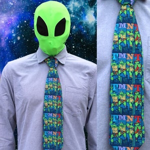 May include: A blue tie with a repeating pattern of Teenage Mutant Ninja Turtles characters and the letters TMNT. The tie is on a light blue shirt with a green alien mask on top.