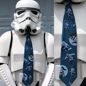 May include: A blue necktie featuring a repeating pattern of cartoon characters in action poses. The tie is worn by a Stormtrooper costume. The characters are in white and gray against a blue background. The tie is a fun accessory.