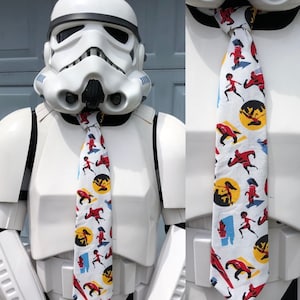 May include: A white stormtrooper action figure wearing a white tie with a colorful pattern of cartoon superheroes. The tie features a repeating pattern of red, yellow, and blue superheroes running and jumping.