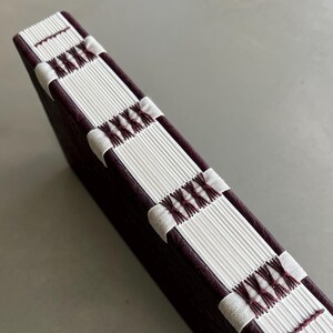 May include: A handmade journal with a dark burgundy leather cover. The spine features white paper sections stitched together with burgundy thread. The journal is angled, showcasing the detailed craftsmanship and texture of the materials.