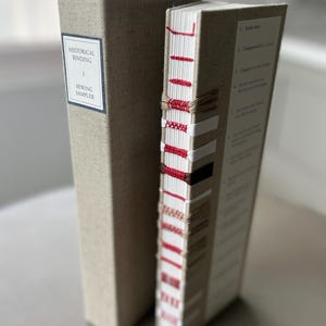 Op de afbeelding: Twee boeken gebonden in linnen stof. Het boek links heeft de tekst "HISTORICAL BINDING + SEWING SAMPLER" op de rug. Het boek rechts heeft een rood en wit gestikte rug met de tekst "K-stitch" op de rug.
