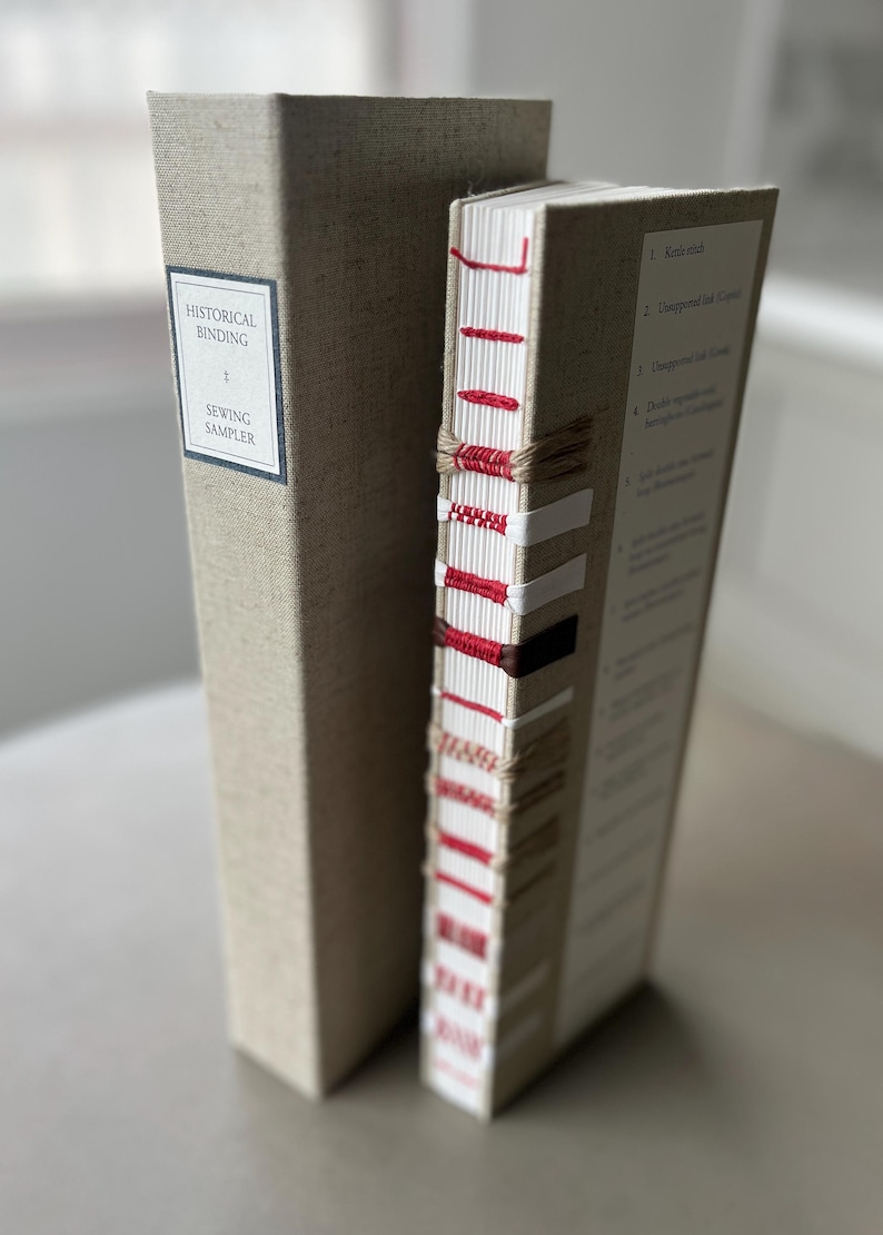 Op de afbeelding: Twee boeken gebonden in linnen stof. Het boek links heeft de tekst "HISTORICAL BINDING + SEWING SAMPLER" op de rug. Het boek rechts heeft een rood en wit gestikte rug met de tekst "K-stitch" op de rug.