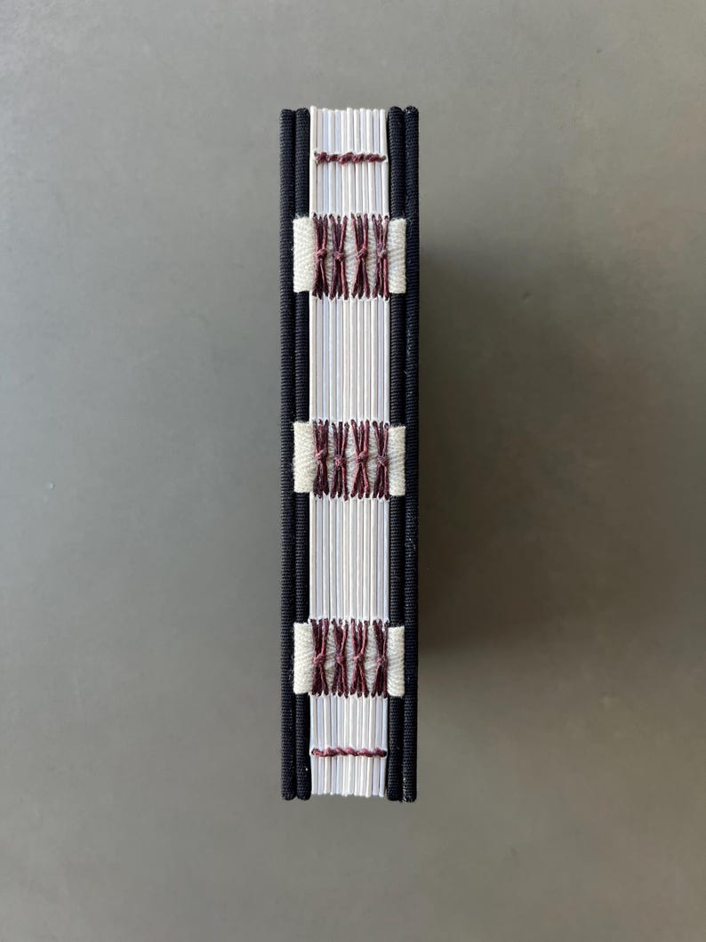 Op de afbeelding: Een klein, wit boek met een zwarte omslag en bruine stiksels. Het boek is gebonden met een koptische steek.