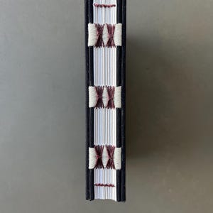 Op de afbeelding: Een klein, wit boek met een zwarte omslag en een rug gebonden met wit en bordeauxrood garen. Het boek is gesloten en staat rechtop.
