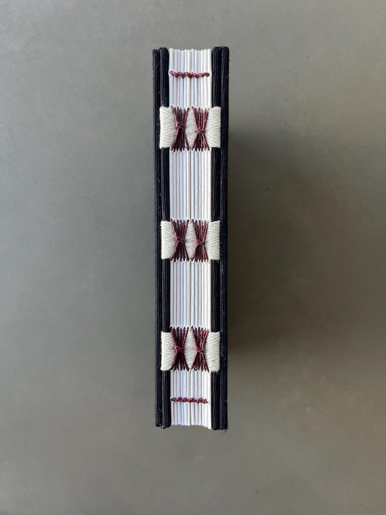 Op de afbeelding: Een klein, wit boek met een zwarte omslag en een rug gebonden met wit en bordeauxrood garen. Het boek is gesloten en staat rechtop.