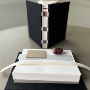 May include: A black bound journal with a stack of white paper, a spool of brown thread, a piece of gray cardstock, and a white ribbon.