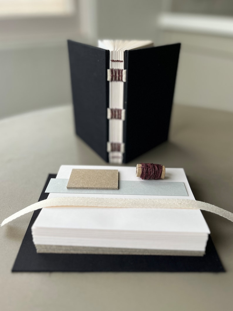 May include: A black bound journal with a stack of white paper, a spool of brown thread, a piece of gray cardstock, and a white ribbon.