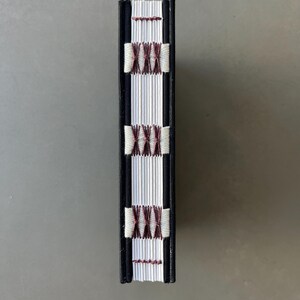 Op de afbeelding: De rug van een klein, zwart gebonden boek met witte pagina's en een decoratief steekpatroon in bordeauxrood en wit.