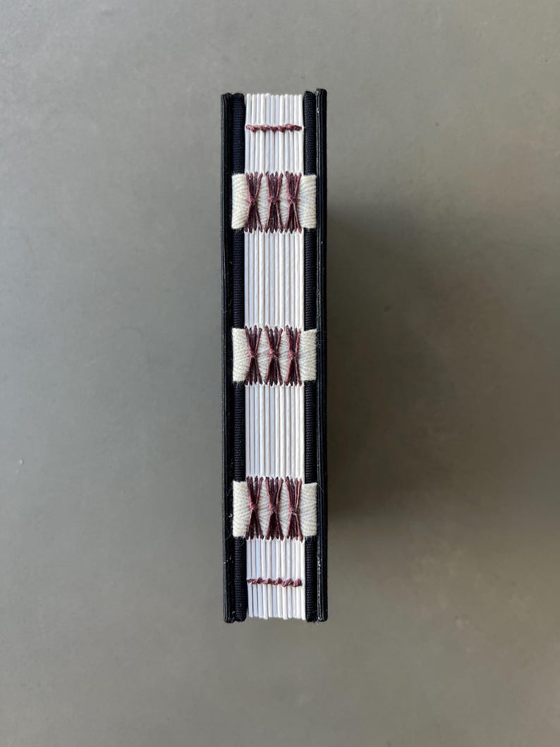 Op de afbeelding: De rug van een klein, zwart gebonden boek met witte pagina's en een decoratief steekpatroon in bordeauxrood en wit.