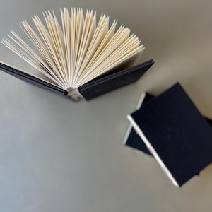 Op de afbeelding: Een zwart hardback-journaal met een gewaaierde stapel blanco pagina's. Het journaal ligt open op een lichtgrijze ondergrond. In de rechterbenedenhoek van de afbeelding liggen nog twee zwarte journalen op elkaar.