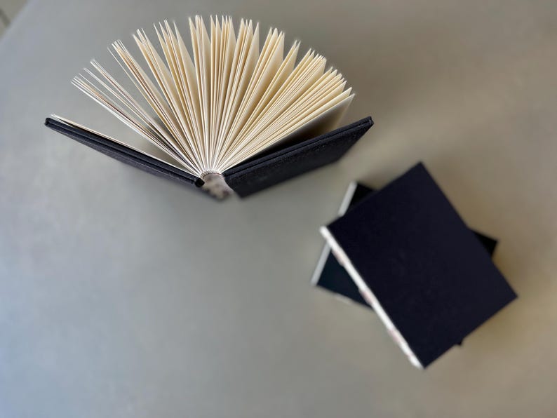 Op de afbeelding: Een zwart hardback-journaal met een gewaaierde stapel blanco pagina's. Het journaal ligt open op een lichtgrijze ondergrond. In de rechterbenedenhoek van de afbeelding liggen nog twee zwarte journalen op elkaar.