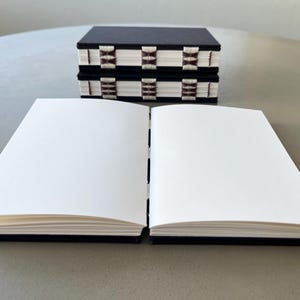 May include: Two black hardcover journals with white and brown patterned spines are stacked on top of each other. A third journal with a black cover is open and lying flat on a gray surface.