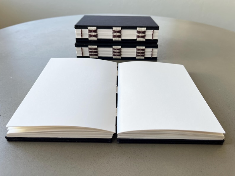 May include: Two black hardcover journals with white and brown patterned spines are stacked on top of each other. A third journal with a black cover is open and lying flat on a gray surface.