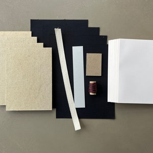 May include: A set of bookbinding supplies including a stack of white paper, a stack of brown paper, black fabric, a white ribbon, a light blue cardstock, a brown cardstock, and a spool of brown thread.