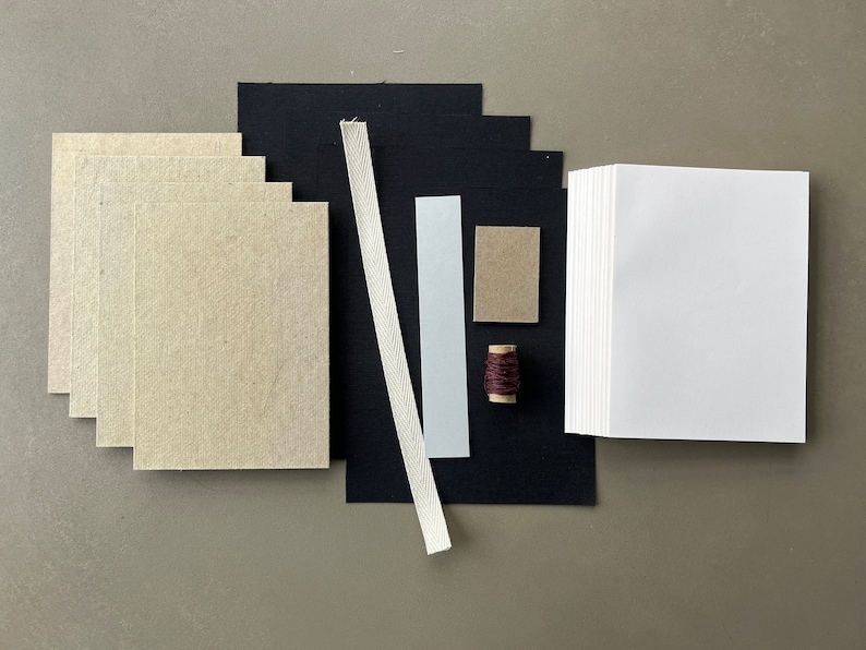 May include: A set of bookbinding supplies including a stack of white paper, a stack of brown paper, black fabric, a white ribbon, a light blue cardstock, a brown cardstock, and a spool of brown thread.