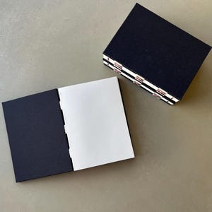 Op de afbeelding: Twee zwarte notitieboekjes met witte pagina's. Het notitieboekje links is open en laat de lege witte pagina's zien. Het notitieboekje rechts is gesloten en gestapeld op een ander notitieboekje.
