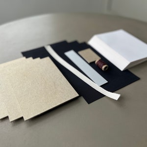 May include: A set of craft supplies for bookbinding, including beige cardstock, black cardstock, white ribbon, a small piece of brown cardstock, and a spool of brown thread.