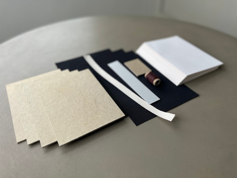 May include: A set of craft supplies for bookbinding, including beige cardstock, black cardstock, white ribbon, a small piece of brown cardstock, and a spool of brown thread.