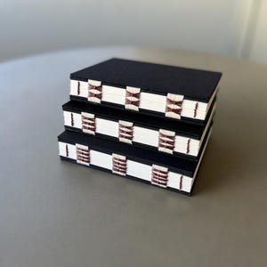 May include: Three black notebooks stacked on top of each other. The notebooks have white covers with a burgundy and white patterned design.