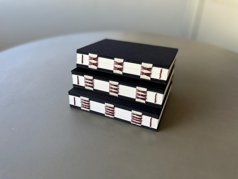 May include: Three black notebooks stacked on top of each other. The notebooks have white covers with a burgundy and white patterned design.