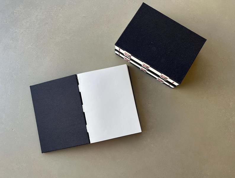May include: Two black notebooks with white pages. The notebook on the left is open, showing the blank white pages. The notebook on the right is closed and stacked on top of another notebook.