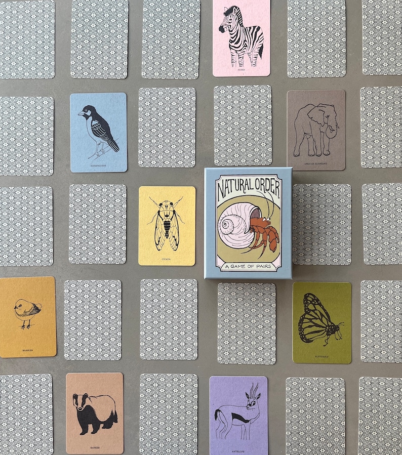 May include: A set of cards featuring illustrations of various animals and insects, including a zebra, elephant, cicada, butterfly, and more. The cards are arranged in a grid pattern on a gray surface. The box for the game is in the center of the image and says "Natural Order: A Game of Pairs".