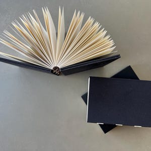 May include: Two black hardcover journals with blank pages. The journal on the left is open, revealing the blank pages inside. The journal on the right is closed.