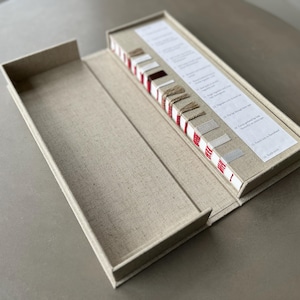 Op de afbeelding: Een gesloten, met linnen beklede doos met een rood-wit gestreept boek met stofstalen erin. Het boek bevat verschillende stofstalen en beschrijvingen.